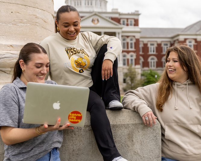 three mizzou students stand in front of jesse hall by the columns laughing and looking at a laptop
