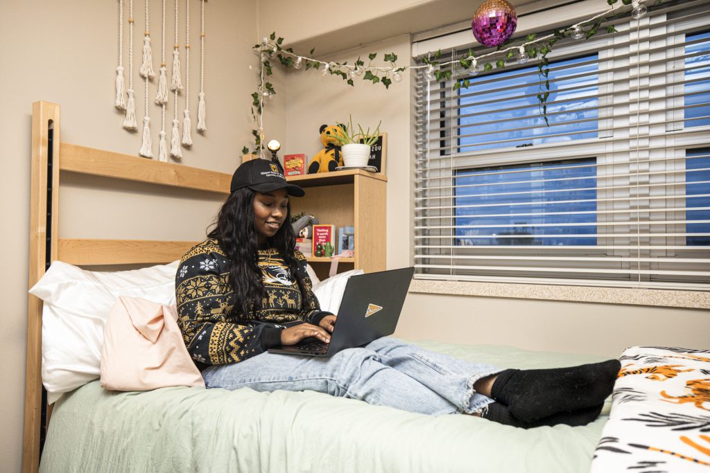 Mizzou student sitting on her bed in a mizzou sweatshirt and using her laptop