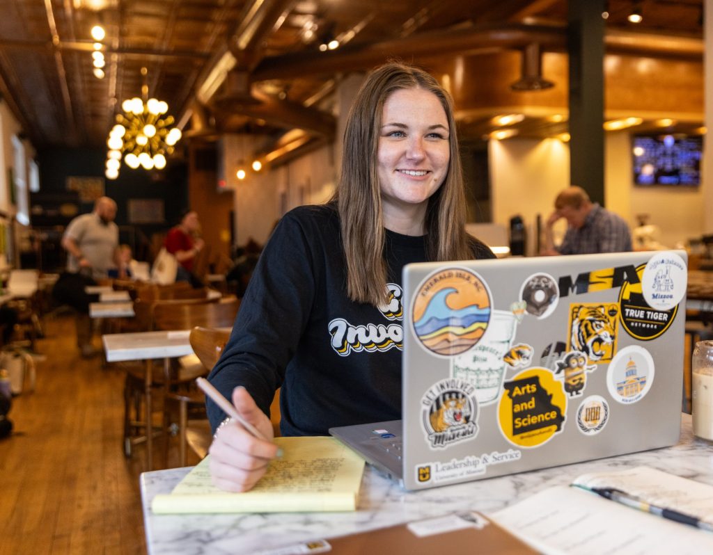 Mizzou student studying in a coffeeshop with their laptop open and covered in Mizzou stickers